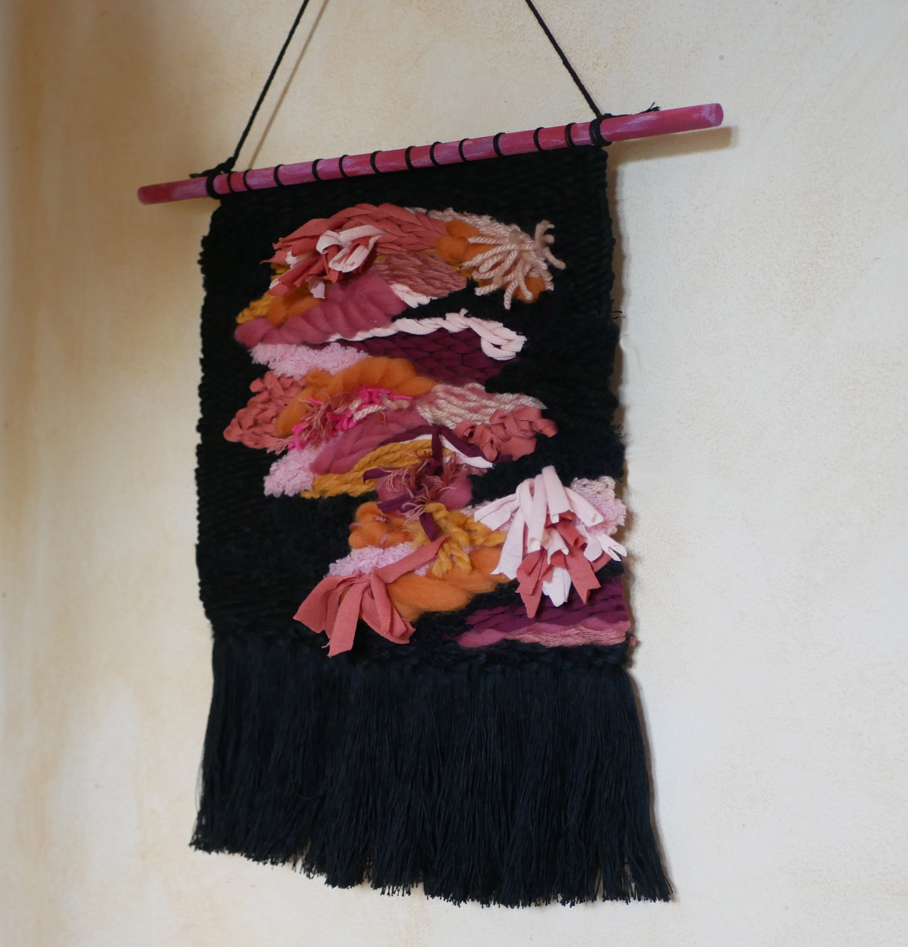 Suspension tissage mural d'inspiration florale en laines et coton de couleurs roses sur noir aux longues franges