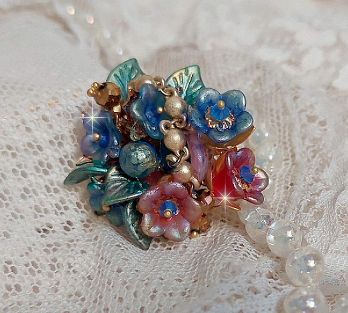 Bague Bel Air montée avec des fleurs lucites peintes à la main aux couleurs : Bleu, Rose, Rouge, Argent, Blanc avec feuilles ondulées Vert Cuivré à laquelle s’ajoute perles Dorées en Verre