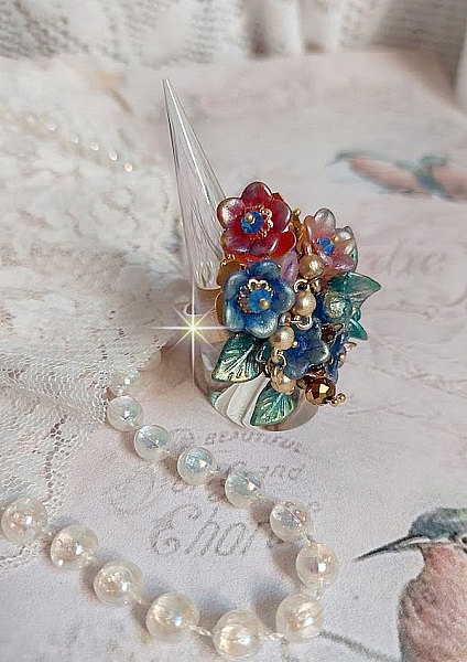 Bague Bel Air mont&eacute;e avec des fleurs lucites peintes &agrave; la main aux couleurs : Bleu, Rose, Rouge, Argent, Blanc avec feuilles ondul&eacute;es Vert Cuivr&eacute; &agrave; laquelle s&rsquo;ajoute perles Dor&eacute;es en Verre