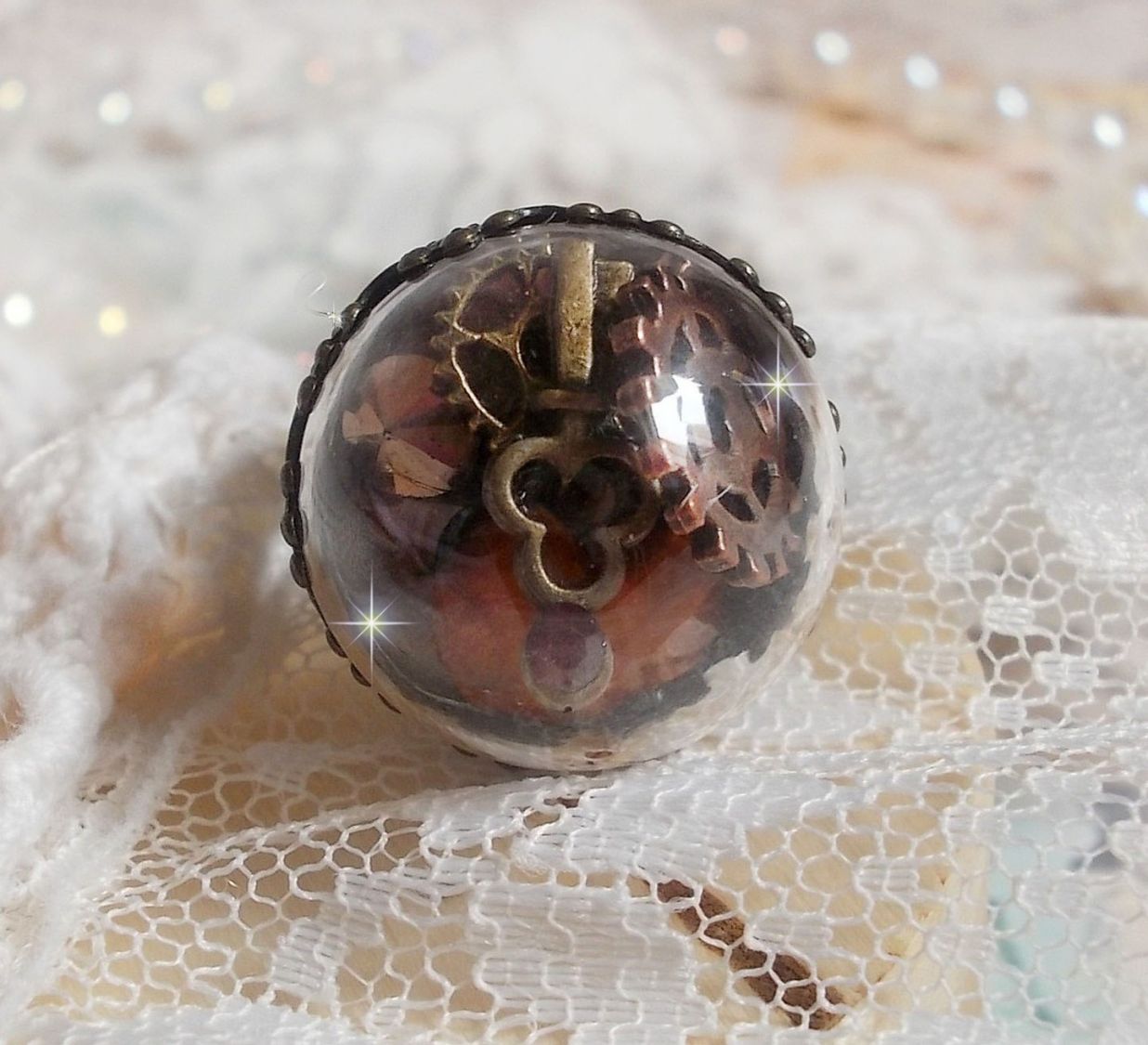 Bague Majesté Steampunk aux rouages mécaniques à laquelle s’ajoute une clé et un strass de couleur Siam et un strass en verre Doré. Le tout est mis sous un globe en verre sur une bague réglable laiton