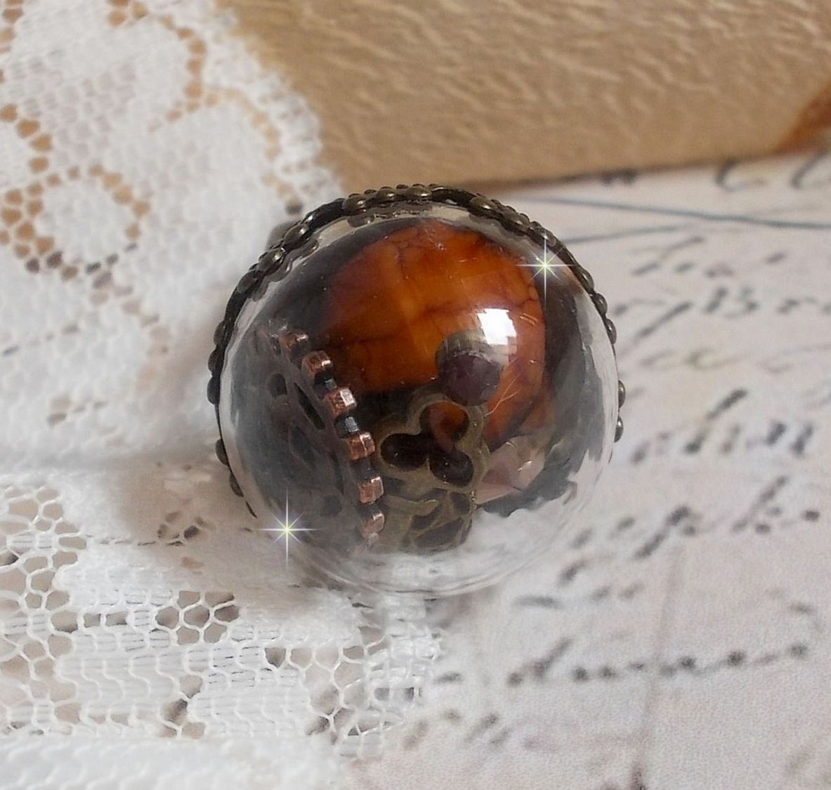 Bague Majesté Steampunk aux rouages mécaniques à laquelle s’ajoute une clé et un strass de couleur Siam et un strass en verre Doré. Le tout est mis sous un globe en verre sur une bague réglable laiton