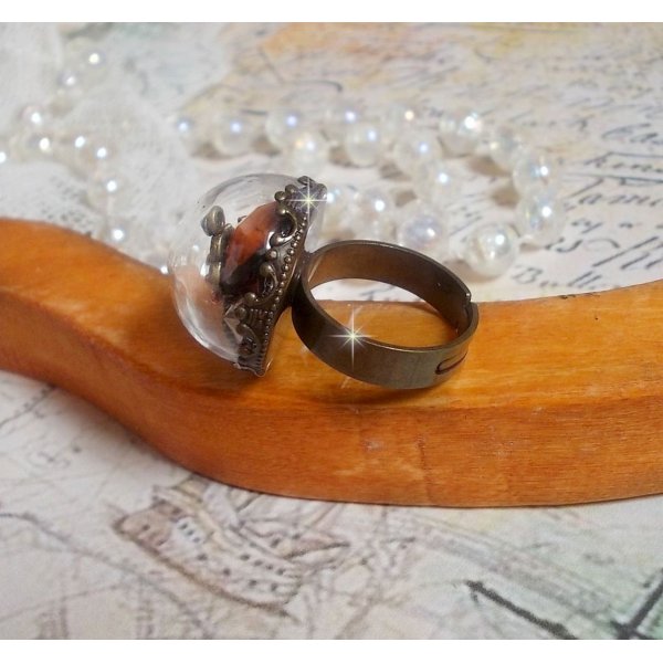 Bague Majest&eacute; Steampunk aux rouages m&eacute;caniques &agrave; laquelle s&rsquo;ajoute une cl&eacute; et un strass de couleur Siam et un strass en verre Dor&eacute;. Le tout est mis sous un globe en verre sur une bague r&eacute;glable laiton