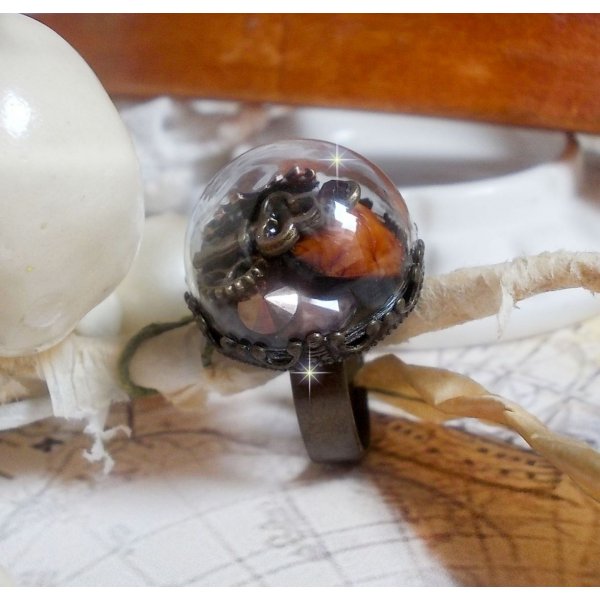 Bague Majest&eacute; Steampunk aux rouages m&eacute;caniques &agrave; laquelle s&rsquo;ajoute une cl&eacute; et un strass de couleur Siam et un strass en verre Dor&eacute;. Le tout est mis sous un globe en verre sur une bague r&eacute;glable laiton