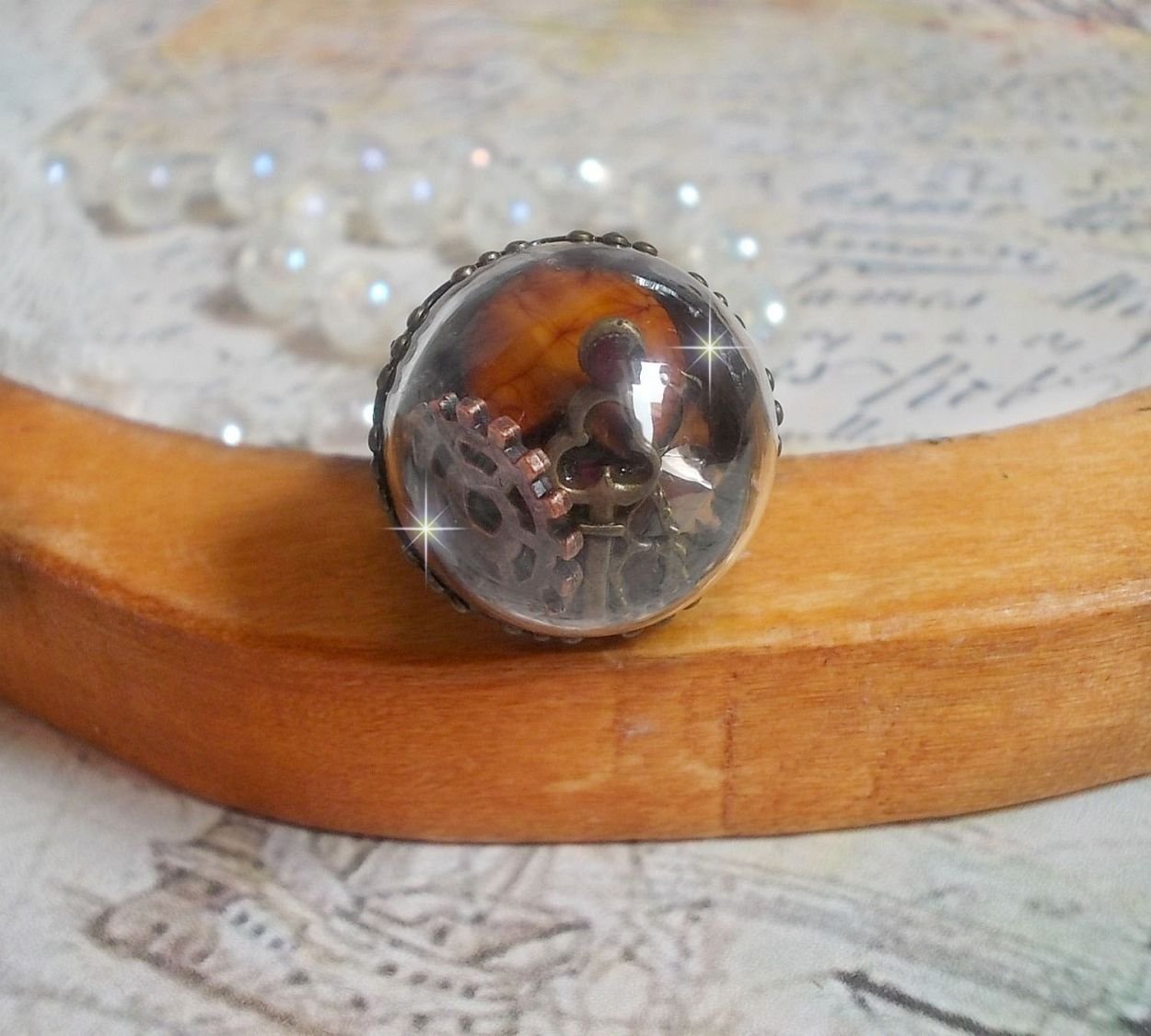 Bague Majesté Steampunk aux rouages mécaniques à laquelle s’ajoute une clé et un strass de couleur Siam et un strass en verre Doré. Le tout est mis sous un globe en verre sur une bague réglable laiton