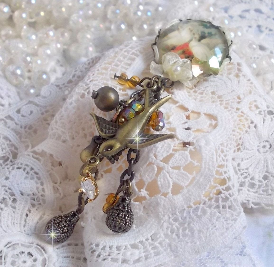 Broche Colombes aux plumages blancs avec des breloques filigranées gouttes et lisses, perles rondes et nœud en cristal, ruban organza crème, pendentif plaqué or avec un zircon sur une broche en métal