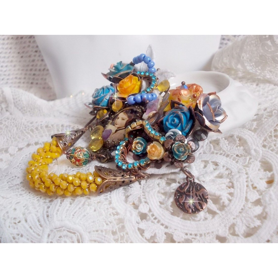 Broche Eclats Pétillants d’inspiration Baroque, ornée de fleurs en résine autour d’un cabochon strassés et perlés sur estampes, cristaux, breloques, perle cloisonnée, perles jaunes en verre et roses. 