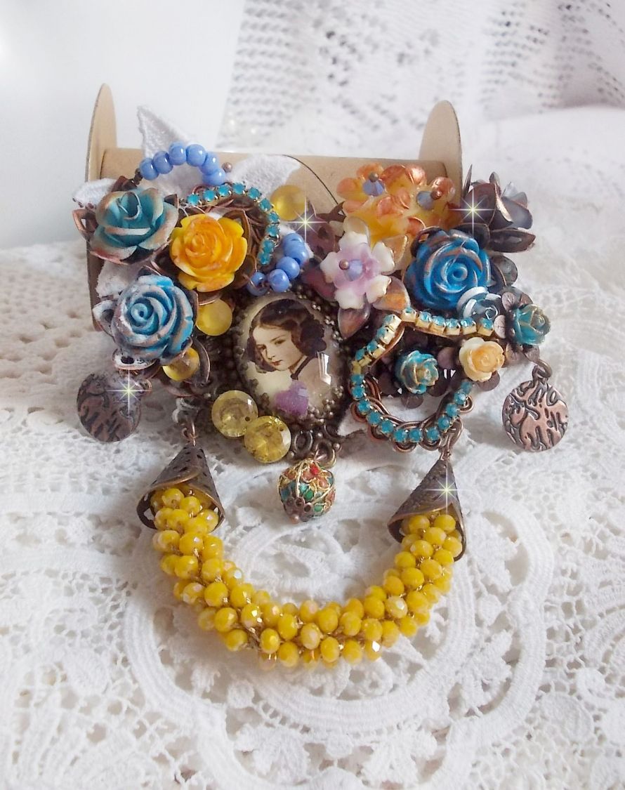 Broche Eclats Pétillants d’inspiration Baroque, ornée de fleurs en résine autour d’un cabochon strassés et perlés sur estampes, cristaux, breloques, perle cloisonnée, perles jaunes en verre et roses. 