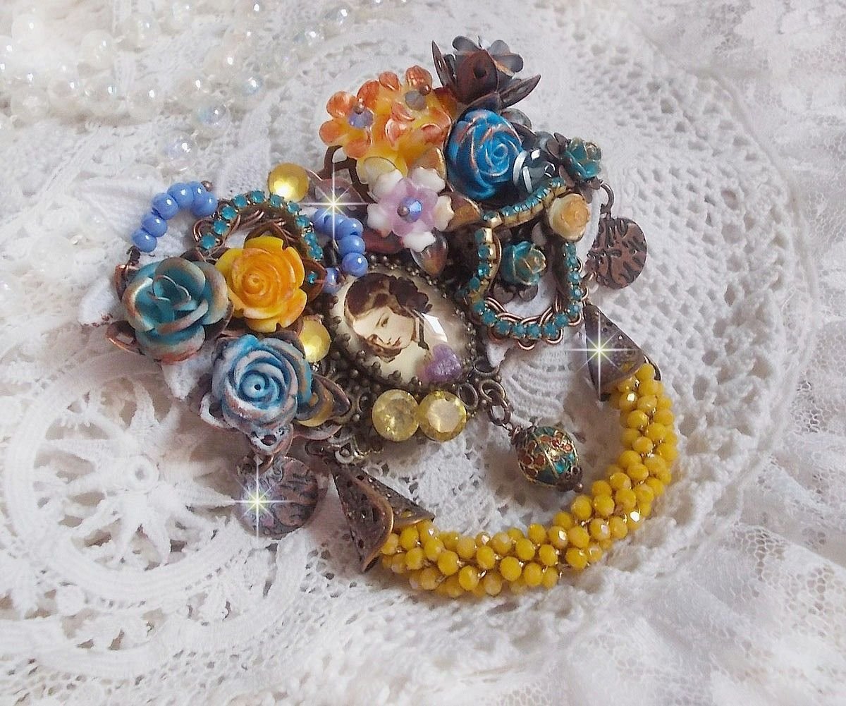 Broche Eclats Pétillants d’inspiration Baroque, ornée de fleurs en résine autour d’un cabochon strassés et perlés sur estampes, cristaux, breloques, perle cloisonnée, perles jaunes en verre et roses. 