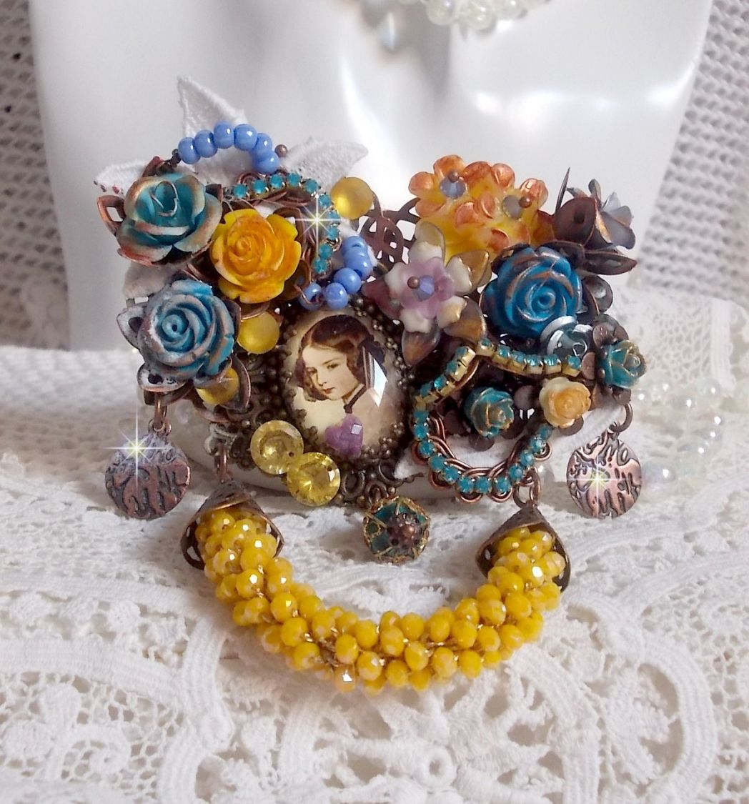 Broche Eclats Pétillants d’inspiration Baroque, ornée de fleurs en résine autour d’un cabochon strassés et perlés sur estampes, cristaux, breloques, perle cloisonnée, perles jaunes en verre et roses. 